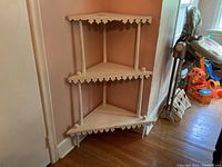 Full view of vintage corner shelf with three shelves and decorative carved trim along edges. Light cream paint with visible wear and damage to edges.
