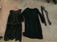 Two black vintage flapper style dresses on hangers laid on the floor along with two belts.