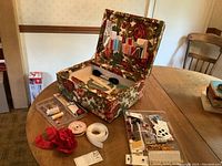 Full view of vintage sewing kit box open showing interior organization with threads, trims, needles, and other notions on a wooden table.