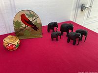 Wide shot of all items displayed on red cloth: five black wooden elephants, one with visible repair, small round covered dish with red flower handpaint, and semi-circular stand with handpainted red bird and leaves.