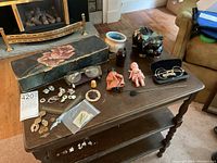 Overview of table with military buttons, badges, vintage dolls, piggy bank, eyeglasses, stamps, large floral box, and other collectibles arranged for inspection.