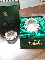 Two Belleek boxes displayed, one open showing the oval lace basket with flower accents, and the other with the smaller round basket beside it.