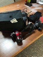 Lot overview showing red Nikon Coolpix P510 with strap, black camera bag, charger, cord, and black Tasco binoculars with case on a wooden surface.