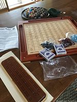 Photo showing wooden One-Eyed-Jack game board with printed grid, bags containing black and red gaming chips, two decks of playing cards, wooden cribbage board with pegs, and printed instructions.