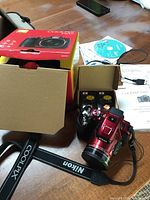 Red Nikon Coolpix P610 camera with original box, strap, charger, cord, manual and software CD on table