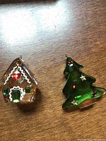Both Swarovski crystal Christmas ornaments, gingerbread house and green tree, shown on wooden surface.