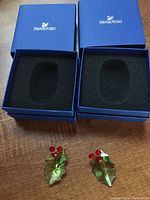 Two green crystal holly leaf Christmas ornaments with red berries shown outside their blue Swarovski boxes with foam inserts on a wooden surface.