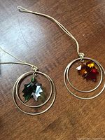 Two Swarovski crystal ornaments on wood surface, each with a layered silver-tone ring and hanging crystal leaf in autumnal colors and green.