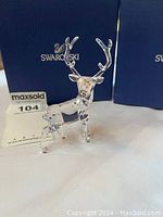 Side angled view of the clear Swarovski crystal stag figurine with faceted body and detailed antlers, displayed in front of blue Swarovski boxes.