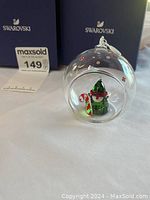 Clear glass ball Christmas ornament with Swarovski elf figure inside on white surface with two Swarovski boxes in background and a MaxSold bid number.