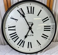 Full front view of large black framed round wall clock with Roman numerals and black hands, reading UNION STATION No.570.