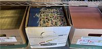 Three cardboard boxes filled with scrapbook paper sheets arranged on a metal shelving unit.