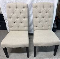Two beige fabric chairs side by side with pleated button tufted high backs and dark wooden legs.