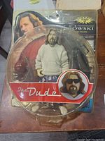 Front view of The Dude collectible action figure inside original blister packaging. The figure wears a grey hoodie and pajama pants, holding a red bowling ball. Packaging shows The Dude character with hair and beard, and an inset of the figure's head with sunglasses.