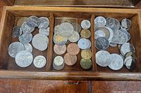 Top-down view inside wooden box showing various old silver, bronze, and copper coins sorted into three compartments.