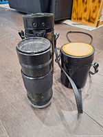 Shows the camera telephoto lens with its attached black cylindrical case, and a second empty black lens case, all placed on a wooden floor.