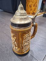 Front angle view of vintage German beer stein showing detailed relief of floral motifs and a figure with a hat, ceramic body with brown and cream glazing, and ornate pewter lid with pointed finial.