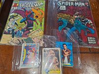Overview of two Spider-Man comic books and several pop culture trading cards in protective sleeves on wooden table.