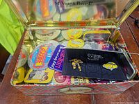 View into the open metal tin showing multiple colorful vintage style buttons and a small black cardboard holder containing three enamel pins.