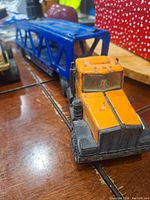 Photo showing the orange cab of the car hauler truck with the blue trailer attached, visible wear and rust.