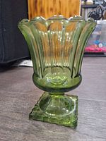 Front view of the green glass vintage vase sitting on floor, showing fluted scalloped rim, vertical ribbing, and square textured base.