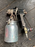 Metal air spray gun lying on wooden surface with metal paint cup attached and air hose fitting connected. Scratches visible on cup and gun body.