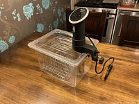 Anova sous vide precision cooker mounted on clear plastic container with dividers on wooden table.