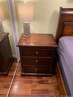 Full view of dark stained three-drawer night table with brass round knob and scalloped base