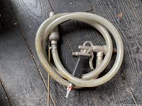 Photo showing air sand blaster with coiled clear reinforced flexible hose, metal nozzle, and control handle on wooden floor.