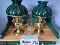 Pair of brass oil lamps with painted green glass shades and a clear glass chimney centered between them on a wooden shelf