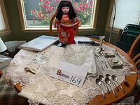 Overall view of table showing linens, silver serving pieces, the silver clock and case, candlestick, and the Japanese doll dressed in red kimono standing upright. Photo shows arrangement and close detail of lace and embroidery on linens.