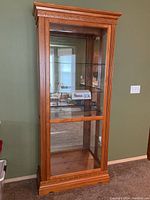 Full front view of the tall wood and glass display cabinet showing carved wood molding, glass front, and shelves inside.