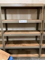 Full front view of shelving unit showing four particle board shelves and the bottom base, all supported by beige/grey perforated metal frame.