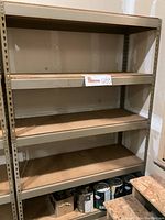 Metal shelving unit with four particle board shelves visible, placed against a wall in a lightly used condition.