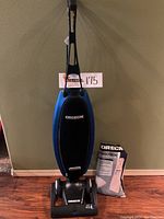 Full view of blue Oreck Magnesium upright vacuum with handle. Next to vacuum is a sealed package of vacuum bags.