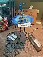 Wide shot showing the metal peacock fountain with iridescent blue glass bowl, the bird feeder pole box, metal plant stands, horseshoes, and thermometers on carpet.