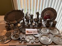Wide view of the entire lot arranged on a table including trays, goblets, candlesticks, spoons, plates, and a red glass vase.