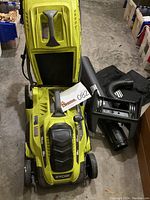 Front view of Ryobi electric mower with top open exposing inner compartment, accessories and grass bag adjacent