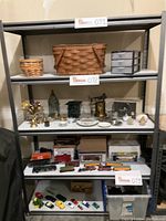 Full view of metal shelving unit showing four white board shelves filled with various items: baskets, small drawers, decorative items, model trains, small toy cars, and boxes underneath.