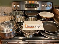Photo showing the full set of stainless steel saucepans, colander, non-stick skillet, pie plate, cake pans, and mixing bowl arranged on stove and countertop.