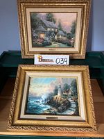 Photo of two framed Thomas Kinkade prints, one depicting a lighted cottage scene, the other a lighthouse coastal landscape. Both have gold-toned ornate frames and cream mats.