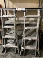Pair of Werner folding aluminum ladders in storage area, folded into step ladder configuration, blue rubber end caps on top hinge.