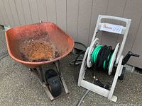 Photo showing an orange metal wheelbarrow and a white hose reel with green garden hose and nozzle.