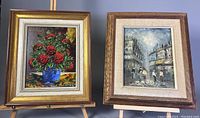 Two framed paintings side by side on easels showing a red rose bouquet and a street scene under illumination.