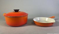 Two orange Le Creuset cast iron pieces, a covered saucepan and a skillet, together in front view