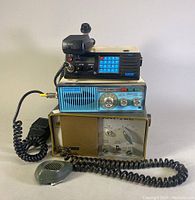 Front view of three stacked vintage marine communication radios, showing the MARINE1 M90 with numeric keypad and microphone on top, the MIDLAND #13-525 radiotelephone in the middle, and RAYTEL TWR-2 CB tube radio on bottom with separate microphone.