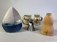 Photo showing the entire lot of five items including the blue and white sailboat ceramic sculpture, two rustic ceramic mugs with handles, the Swedish folk pottery woman figurine holding flowers, and the beige nude torso vase.