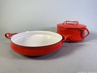 10-inch red enamel cast iron pan with white interior and two looped handles, positioned next to red covered pot
