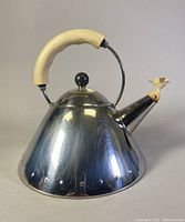 Front and side views of the vintage Alessi stovetop kettle showing stainless steel body and cream-colored handle and bird-shaped whistle.