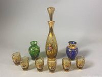 Full set arranged with decanter in center, six shot glasses in front, two colored bud vases behind, showing overall condition and gold gilding.